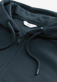 Tumman merensinisen vetoketjullisen hupparin, jossa on narut, pehmeä fleece-vuori, ribattu hihansuu ja kauluksessa lukee "NEXT".