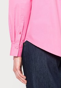 Rosa långärmad skjorta med knappade mudd, med en rundad fåll. Texturen ser slät ut, i kombination med mörkblå denimbyxor.