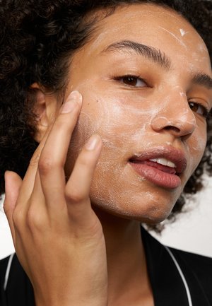 Scrub viso dalla consistenza schiumosa bianca applicato sulla pelle. Il soggetto ha i capelli ricci e uno sfondo neutro. Messa a fuoco sul processo di applicazione.