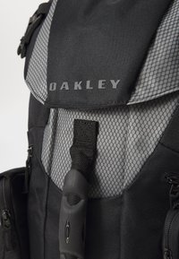 Musta ja harmaa reppu verkkomateriaali yksityiskohdilla, jossa on "Oakley"-logo, säädettävät hihnat ja sileä pintakuvio.