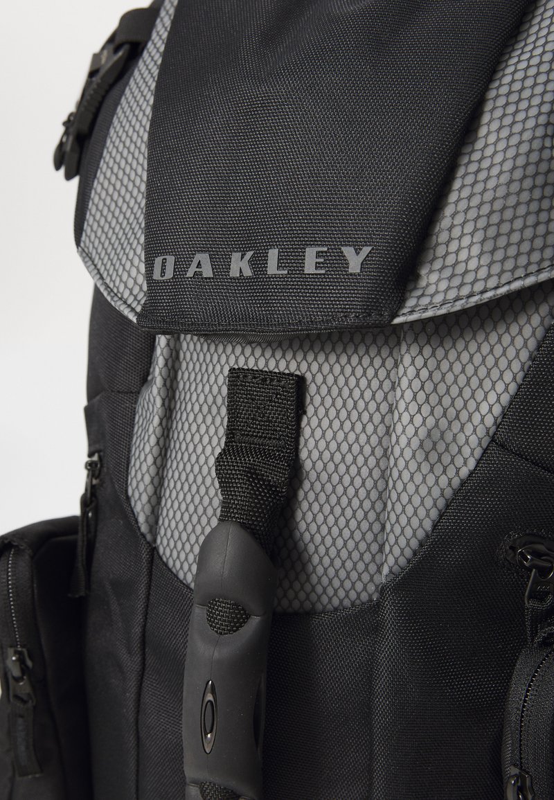 Sac à dos noir et gris avec des accents en maille, arborant le logo "Oakley", des bretelles réglables et une surface lisse.