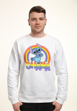 Jonge man met een witte trui met kleurrijke tekst "Ohana", dansend Stitch-personage en een regenboogachtergrond.
