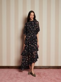Mulher de pé contra uma parede listrada em bege, vestindo um vestido preto com flores e babados, e sapatos pontiagudos de estampa de leopardo, num tapete rosa.