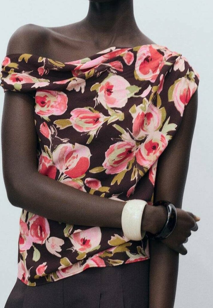 Modèle portant un top marron à fleurs épaules dénudées avec des roses roses, un pantalon sombre, et des bracelets blancs et noirs sur des bras croisés.