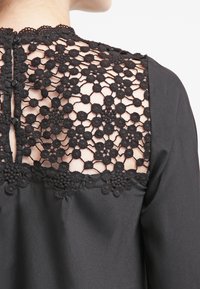Blouse noire avec un dos en dentelle à motifs floraux, fermetures à boutons et une texture satinée lisse à l'avant.