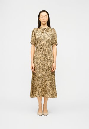 Ženska stoji oblečena v bež midi obleko s črnim abstraktnim potiskom, napihnjenimi rokavi in zavezanim izrezom okoli vratu, kombinirano z bež ostrimi čevlji.