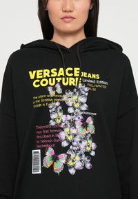 Sudo plano preto com gráficos vibrantes de flores e borboletas. O texto inclui "Versace Jeans Couture" em amarelo. Tecido macio, corte solto.