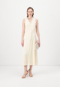Rochie crem fără mâneci, cu accente de franjuri verticale, siluetă în formă de A, material ușor, asortată cu o poșetă bej și sandale cu bretele.