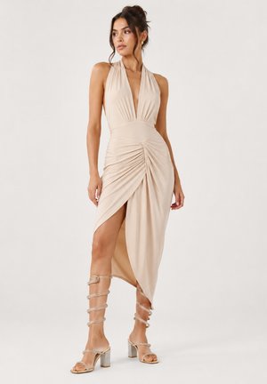Mujer con vestido beige tipo halter fruncido con abertura en el muslo y sandalias doradas de tacón alto con tiras, sobre fondo liso.