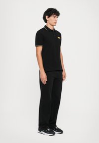 Camisa polo preta com realces amarelos, mangas curtas e um pequeno logótipo no peito, combinada com calças pretas e ténis pretos.