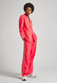 Blazer corallo e pantaloni a gamba larga con strisce laterali dorate, dotati di un solo bottone e tasche. Indossati con sneakers bianche e grigie.