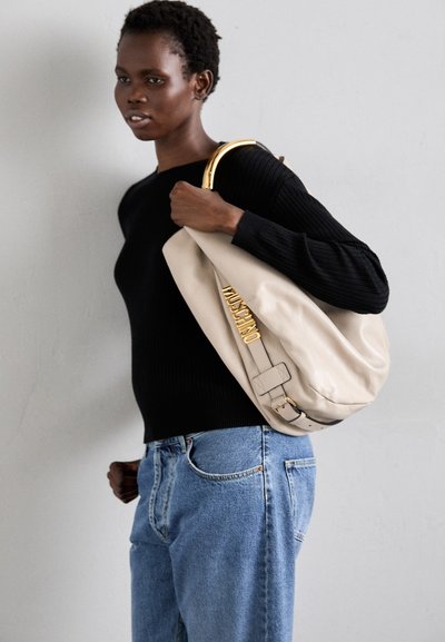 Beige leather shoulder bag with a large, rounded shape, featuring gold hardware and embossed logo detail, paired with a black ribbed sweater and blue jeans.