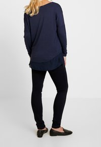 Marinblå långärmad topp med rund nederkant lagrad över en marinblå genomskinlig skjorta, tillsammans med mörka skinny jeans och svarta ballerinaskor, som visar en avslappnad passform.