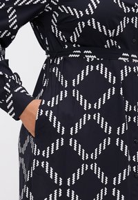 Abito nero con motivo zigzag bianco, dotato di maniche lunghe, vita con cintura e tasche laterali; realizzato in tessuto leggero, chiusura a bottoni.
