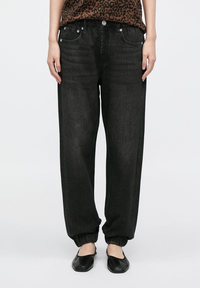 Osoba ubrana w luźne czarne spodnie dresowe z denimu z elastycznymi mankietami, w czarnych, płaskich butach wsuwanych, stojąca na jednolitym tle.