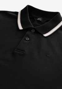 Musta poolopaita, valmistettu teksturoidusta kankaasta, jossa on kaksipainikkeinen aukko, kontrastivärinen raidallinen kaulus ja hillitty kirjailtu logo rinnassa.