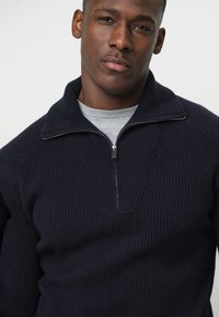 Ciemnobłękitny sweter na zamek o fakturze prążkowanej, z wysokim kołnierzem i głębokim dekoltem w kształcie V. Pod spodem gładka szara koszulka.