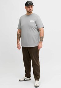 Man with tattoos wearing gray "Jack & Jones" t-shirt, dark brown pants, black cap, and white sneakers standing against a plain white background.