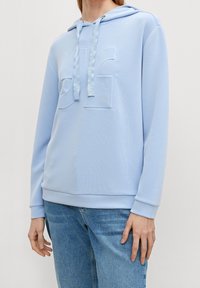 Felpa leggera blu chiaro realizzata in tessuto morbido, con logo in rilievo sulla parte anteriore, cappuccio con cordoncino e polsini e orlo a costine. Abbinata a jeans in denim.