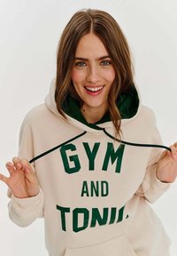 Béžová mikina s kapucí a zelenou podšívkou, se silným zeleným nápisem "GYM AND TONIC". Obsahuje stahovací šňůrku a žebrované manžety.