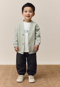 Grijze cardigan met houten knopen, witte T-shirt met autografiek, zwarte sweatpants met elastische boorden, en beige schoenen met klittenbandsluitingen.