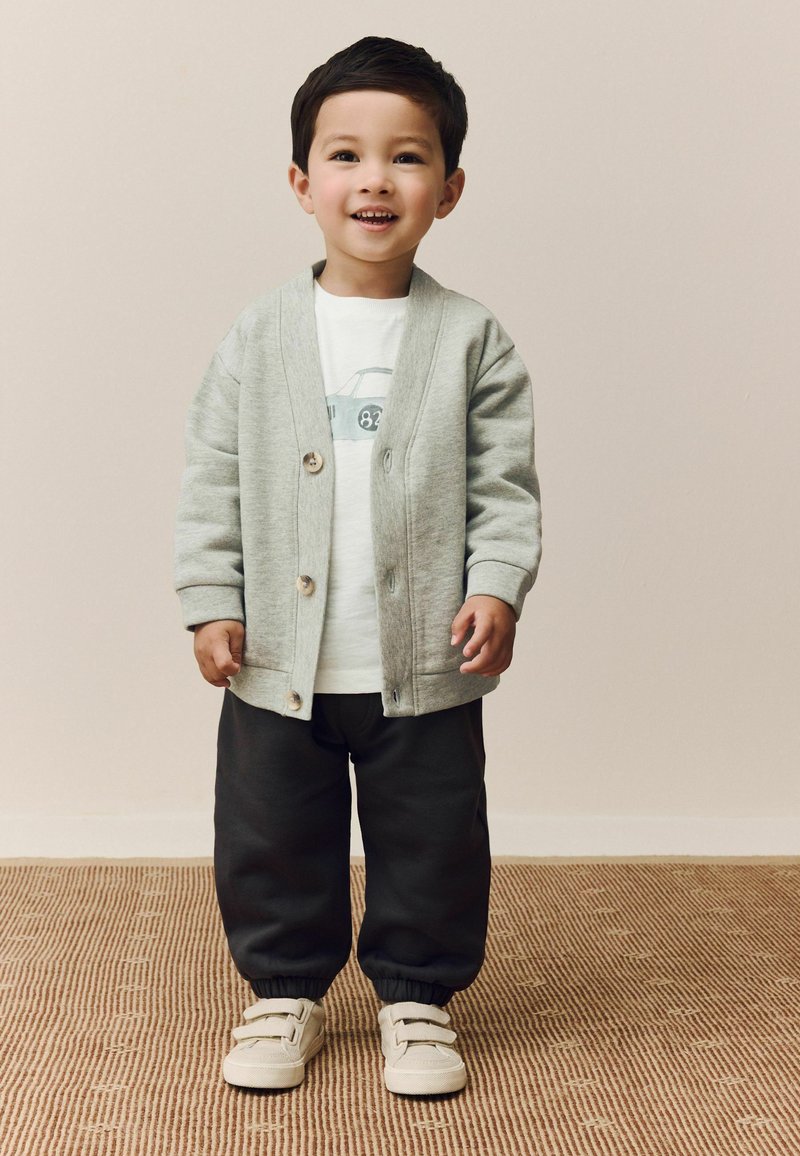 Cardigan grigio con bottoni in legno, T-shirt bianca con grafica di un'auto, pantaloni felpati neri con polsini elasticizzati e scarpe beige con strappi in Velcro.