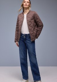 Junge Frau trägt eine braune gesteppte Jacke, ein weißes Hemd, blaue Jeans und weiße Turnschuhe und steht vor einem grauen Hintergrund.