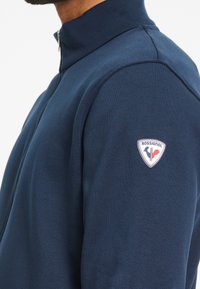 Chaqueta azul marino con cremallera y cuello alto, fabricada en tela suave, con un destacado logo de Rossignol en el hombro izquierdo.