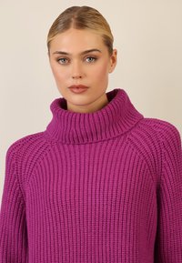 Fioletowy sweter z dzianiny o ribbowym wzorze, z wysokim kołnierzem i luźnym krojem. Posiada wyraźne poziome linie i szerokie rękawy.