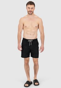 Pantaloni da bagno neri con fascia elastica in vita e cordino bianco. Indossati con sandali neri a ciabatta. Tessuto morbido, design semplice, senza motivi.