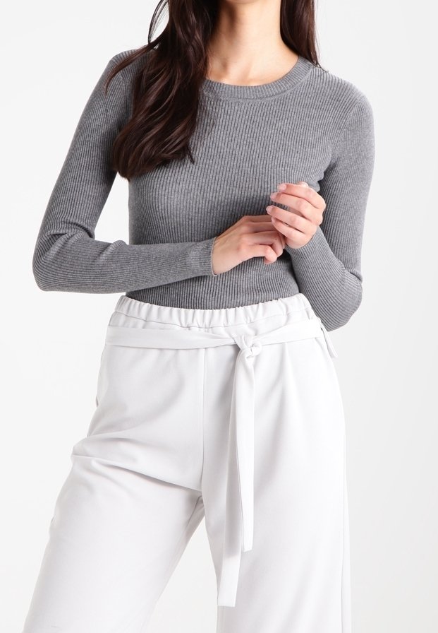 Femme portant un haut gris côtelé à manches longues et ajusté, ainsi qu'un pantalon blanc taille haute avec une ceinture nouée à la taille.