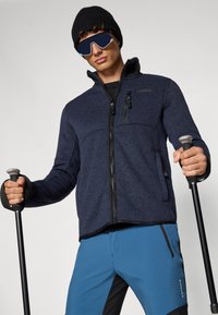 Jachetă din fleece navy cu fermoar frontal, două buzunare cu fermoar și un model texturat, asortată cu pantaloni albaștri și negri și bețe de trekking.
