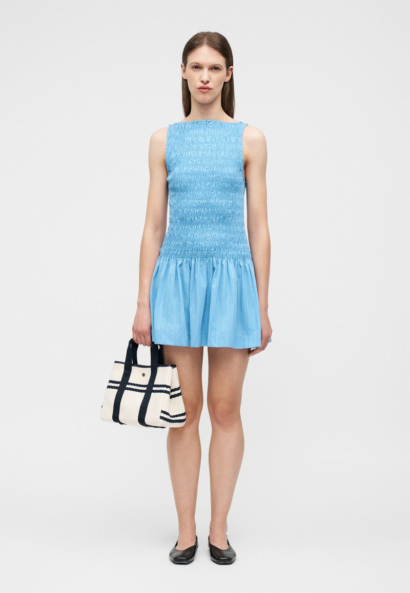 Νεαρή γυναίκα με αμάνικο μπλε φόρεμα, κρατώντας μια τσάντα ώμου με κρεμ και μαύρες ρίγες, φορώντας μαύρα μπαλαρίνες, στέκεται μπροστά σε ένα απλό φόντο.