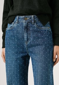 Jeans en denim bleu foncé avec un subtil motif floral blanc. Présente un design standard à cinq poches et une fermeture à bouton en laiton.