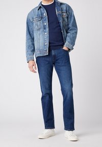 Ljusblå denimjacka med knappstängning och bröstfickor, marinblå tröja under, i kombination med mörkblå jeans och vita sneakers.