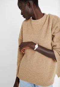 Beige gerippter Strickpullover mit rundem Ausschnitt und seitlichen Schlitzen, kombiniert mit einer hellen Uhr mit rundem Zifferblatt und Lederarmband.