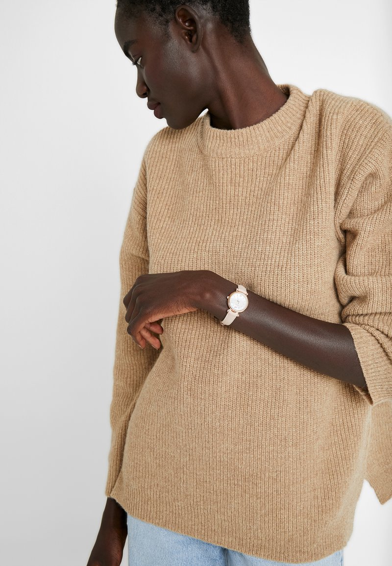 Beige gerippter Strickpullover mit rundem Ausschnitt und seitlichen Schlitzen, kombiniert mit einer hellen Uhr mit rundem Zifferblatt und Lederarmband.