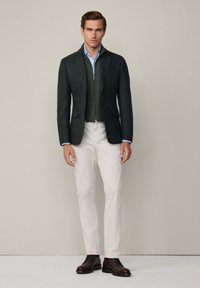 Mörkgrön kavaj över en grön zippered väst, ljusblå skjorta, vita byxor och bruna läderskor, poserande mot en neutral bakgrund.