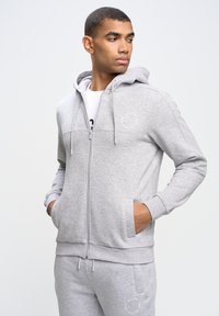 Sudadera con cremallera de color gris claro, hecha de un tejido suave, que presenta un bolsillo tipo canguro, capucha con cordón y un detalle del logo mínimo en el pecho.