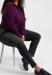 Lila långärmad skjorta med krage som knäpps, i kombination med mörkgrå smala jeans och svarta loafers, sittande på en ljusgrå stol.