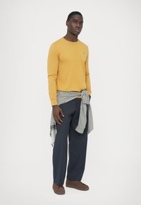 Gelbes Langarm-T-Shirt, dunkelblaue weite Hose, braune Turnschuhe und ein schwarz-weiß kariertes Hemd, das um die Taille gebunden ist.