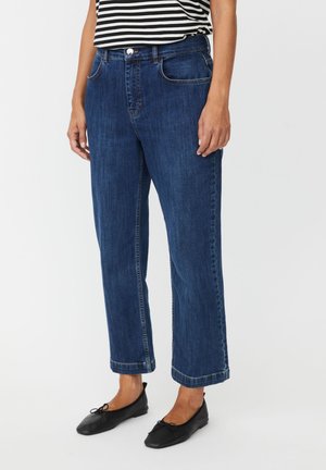 Femme portant un jean droit bleu foncé, des ballerines noires et un haut rayé noir et blanc, debout devant un fond uni.