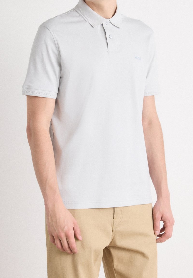 Homme portant un polo gris clair à manches courtes et un pantalon beige, debout devant un fond blanc uni.