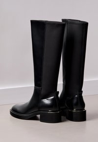 Črni škornji do kolen z gladkim usnjenim sprednjim delom, stranicami iz raztegljive tkanine in masivnim petnim delom z metalnim poudarkom na podplatu.