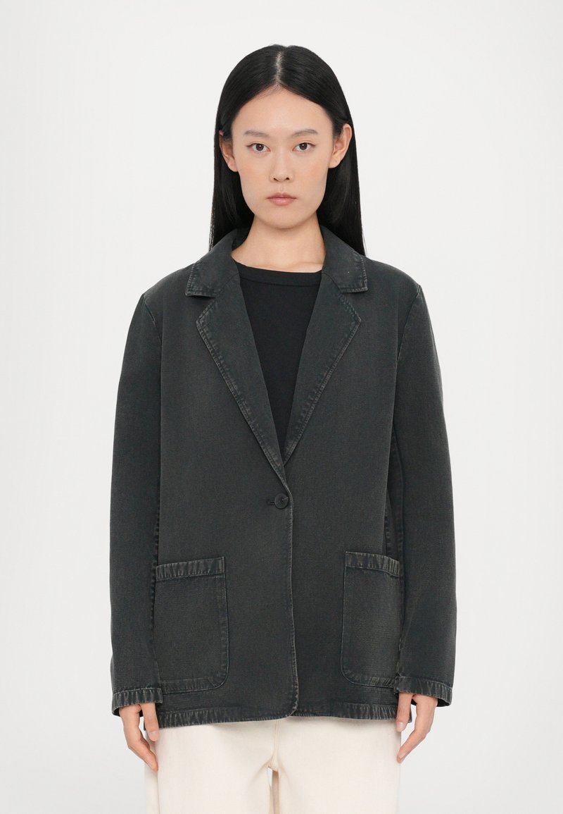 Blazer in denim nero con revers, chiusura a un bottone, due tasche frontali e dettagli di cucitura a vista. Tessuto morbido, vestibilità rilassata.