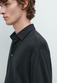 Giovane uomo con capelli ricci marroni che indossa una camicia nera con colletto, mostrato di profilo su uno sfondo uniforme e chiaro.