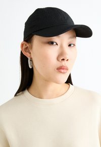 Svart sträv hatt med böjd skärm, bärs med en ljusbeige sweatshirt och cirkulära silverfärgad hoops i öronen.