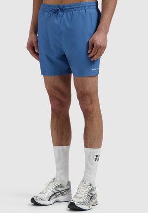 Man wearing blue drawstring shorts, white crew socks with text, and silver running shoes, standing against a plain light background.