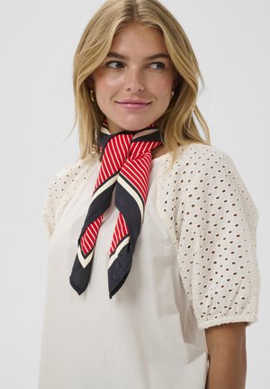 Blonde vrouw die een witte blouse met ooglet-pofmouwen draagt en een rood, zwart-wit gestreepte sjaal om haar nek heeft gestrikt.