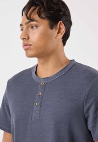 Maglietta henley in cotone blu scuro con maniche corte, texture a coste e tre bottoni di legno sul collo. Design con collo arrotondato.